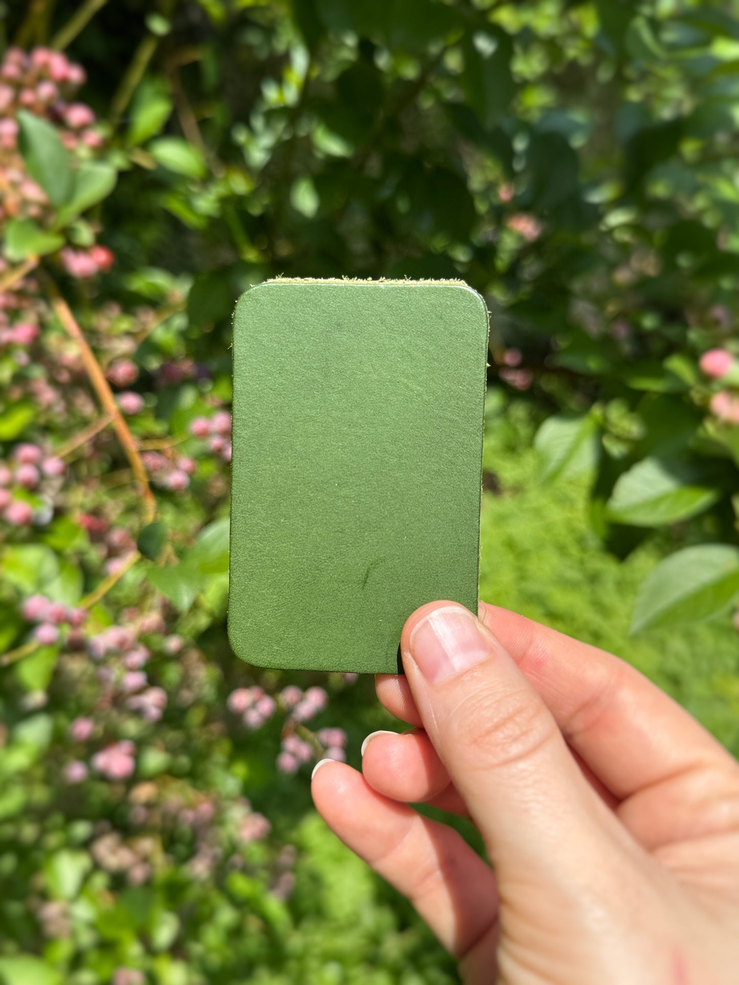 Hand holding a green square leather sample in front of a blurred natural background with greenery and pink flowers.