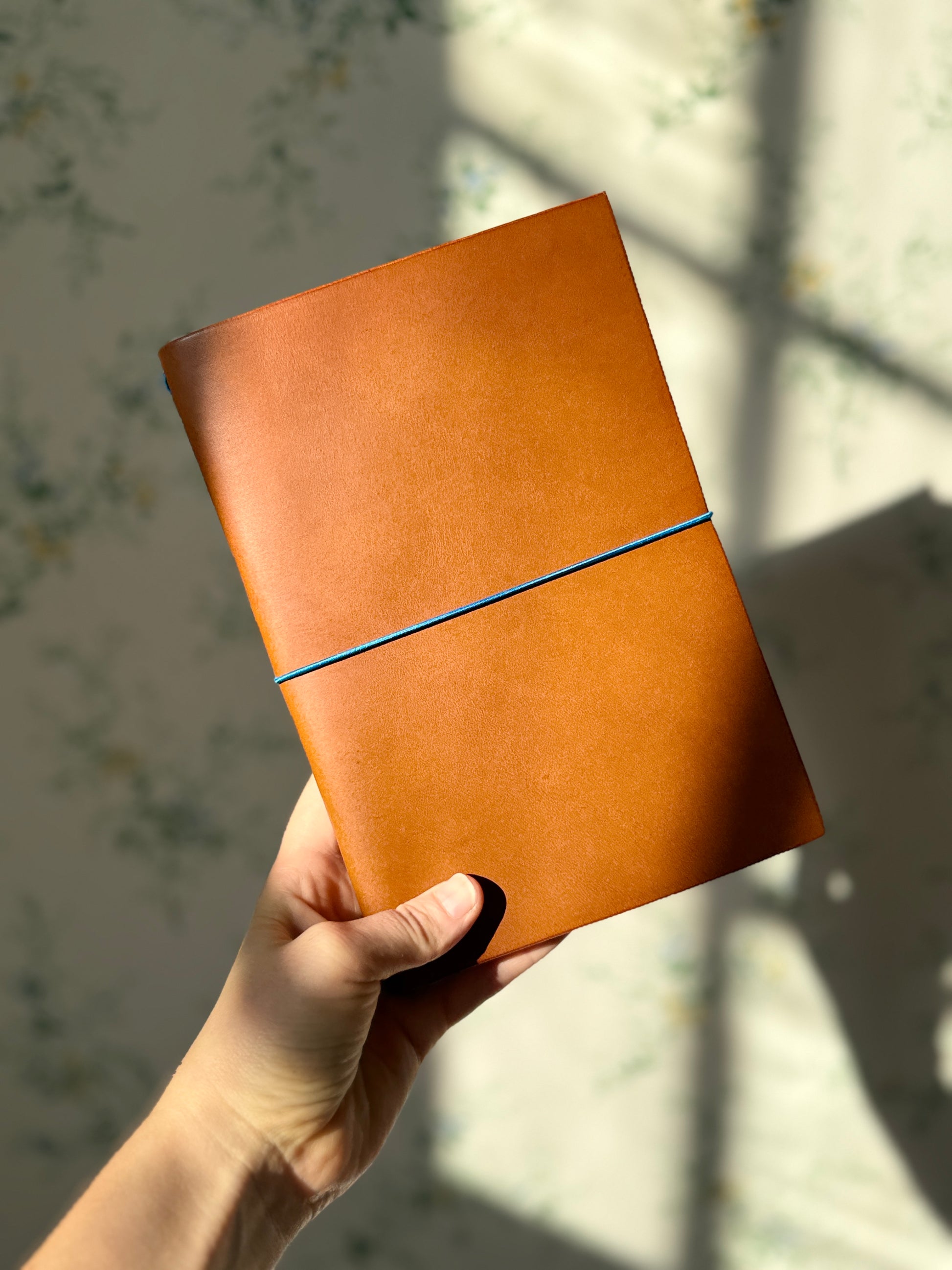 Hand holding a brown leather notebook with a blue strap against a blurred background